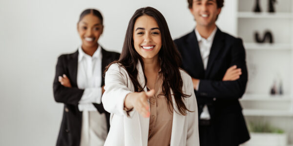 Smiling latin businesswoman HR stretching hand for handshake, greeting to camera, standing with business team indoors